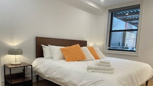 Un lit bien fait, avec du linge de lit blanc et des oreillers orange, une table de chevet avec une lampe, et une fenêtre donnant sur l’extérieur.