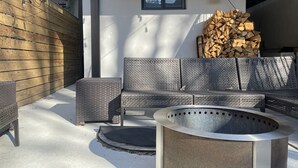 Un espace extérieur aménagé avec un foyer en métal, des meubles en osier et une pile de bois de chauffage adossée à un mur blanc.