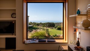 Una habitación con un ventanal grande que da a un paisaje pintoresco, un televisor de pantalla plana, una mesita con una botella y vasos, y una estantería con adornos.