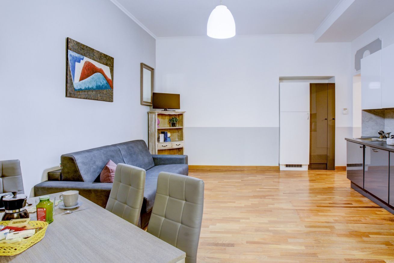 A modern living room with a sofa, dining table, and chairs. There is a kitchen area with cabinets and a countertop. A painting is on the wall, and a television is on a shelf.