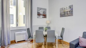 Un coin repas avec une table blanche, quatre chaises grises, un radiateur blanc et une fenêtre avec des rideaux gris.