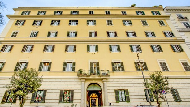 Un immeuble à plusieurs étages, doté d’une façade jaune, de nombreuses fenêtres avec volets et d’une entrée centrale surmontée d’une arche.