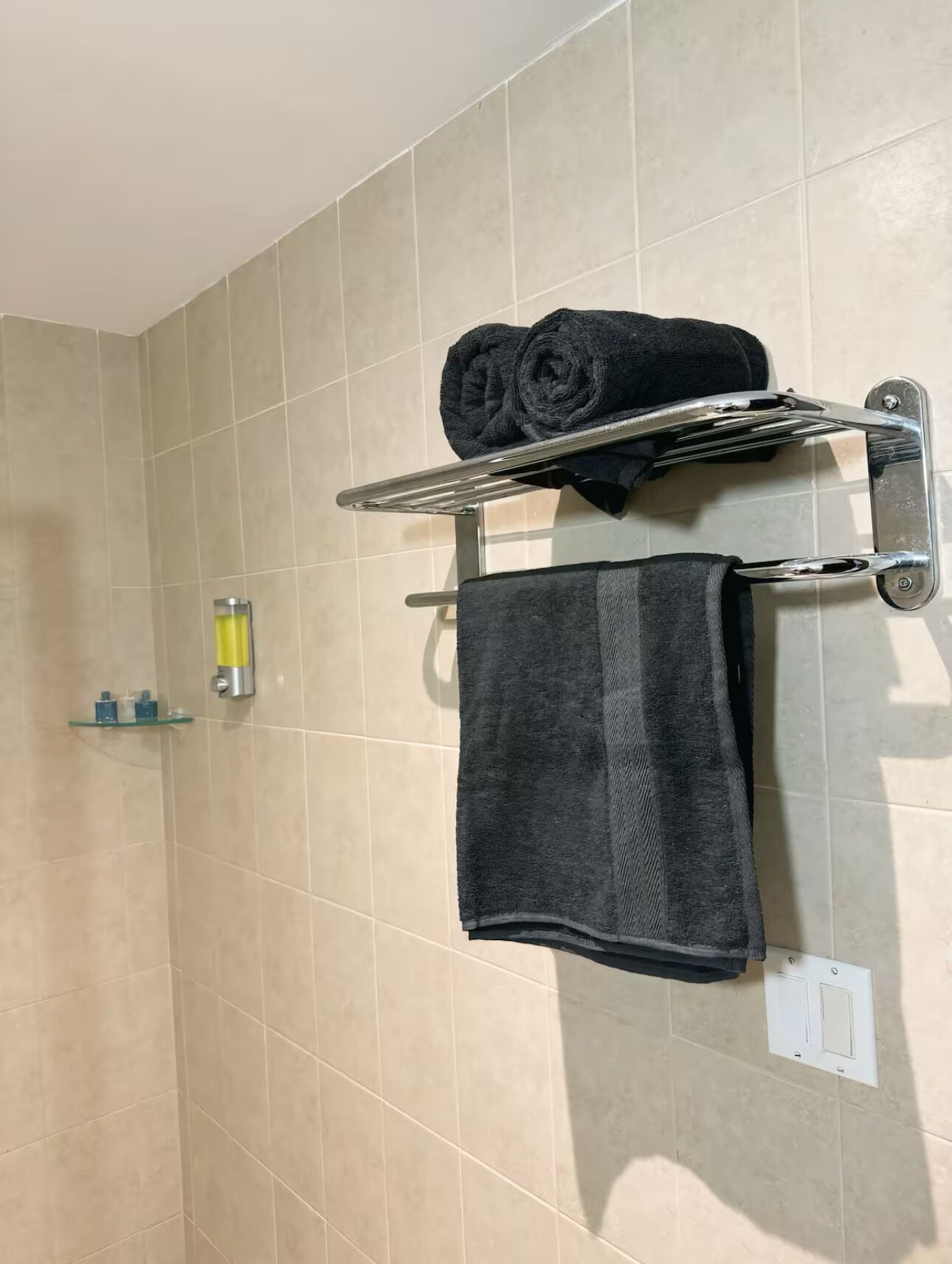 Un baño con baldosas color beige, un perchero con dos toallas y un estante pequeño con artículos de aseo.