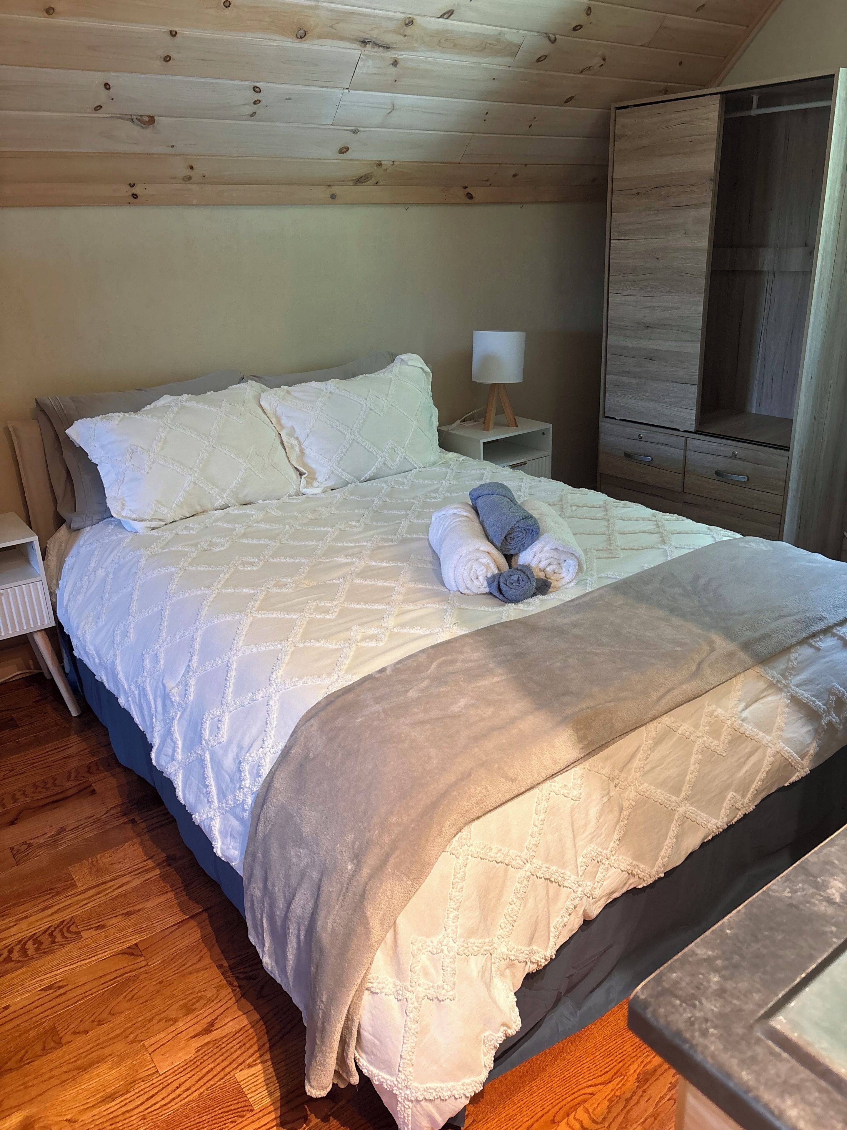 A neatly made bed with a quilt, pillows, and rolled towels. There is a bedside table with a lamp, a wooden wardrobe, and a radiator.