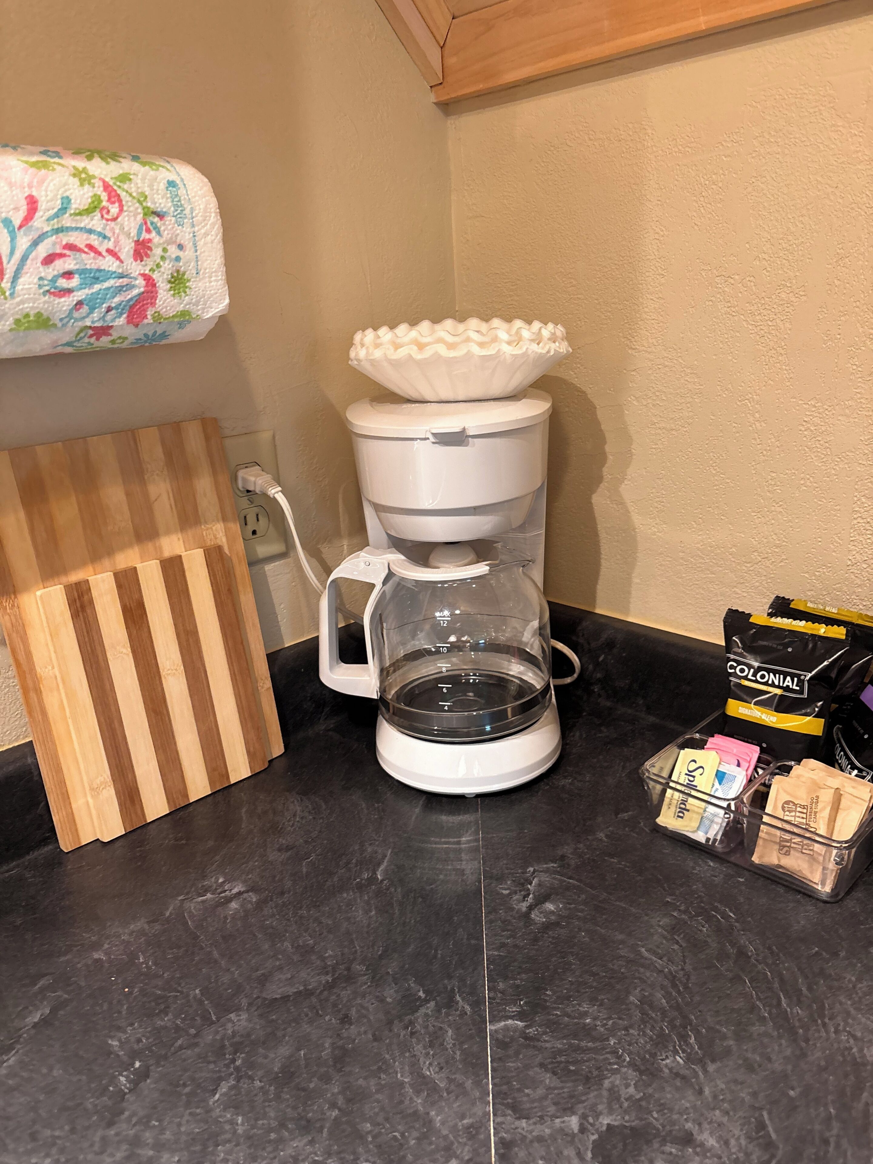 Una macchina da caffè su un piano di lavoro, un tagliere, una confezione di capsule di caffè e un oggetto decorativo al muro.