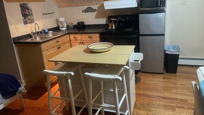 A compact kitchen with wooden cabinets, a microwave, a refrigerator, and a small dining area with two white chairs.