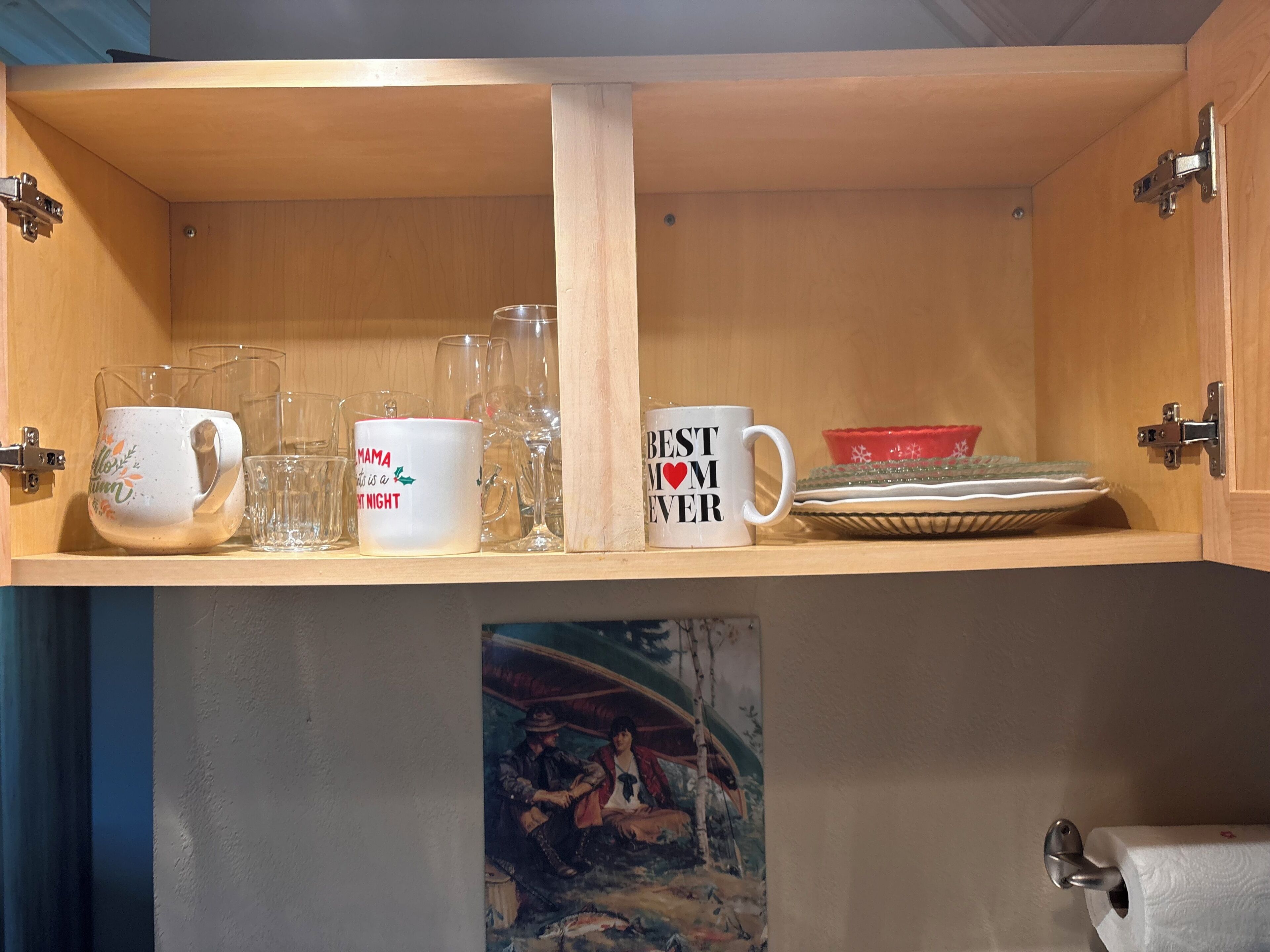 A wooden cabinet with two shelves, one holding clear glassware and a white mug with text, and the other holding a red bowl and a white mug with text.