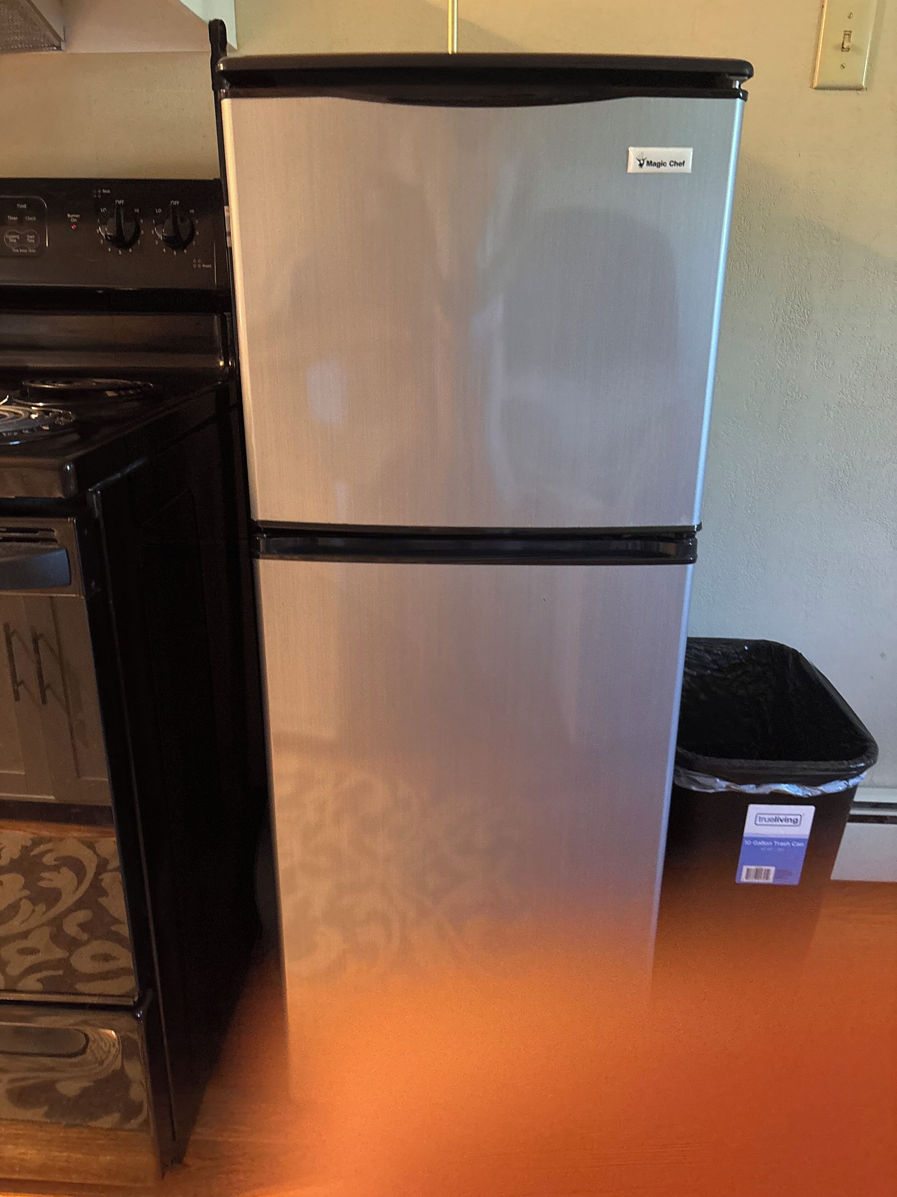 A stainless steel refrigerator with a black base, a trash bin, and a stove in the background.