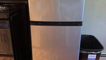 A stainless steel refrigerator with a black base, a trash bin, and a stove in the background.