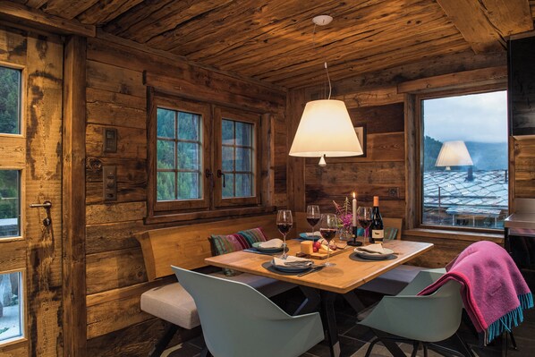 A wooden cabin interior with a dining table set for two, a bench, and a view of a snowy landscape through large windows. - Gmachje - a cozy Tiny House in Findeln with ski-in/ski-out access (Zermatt)