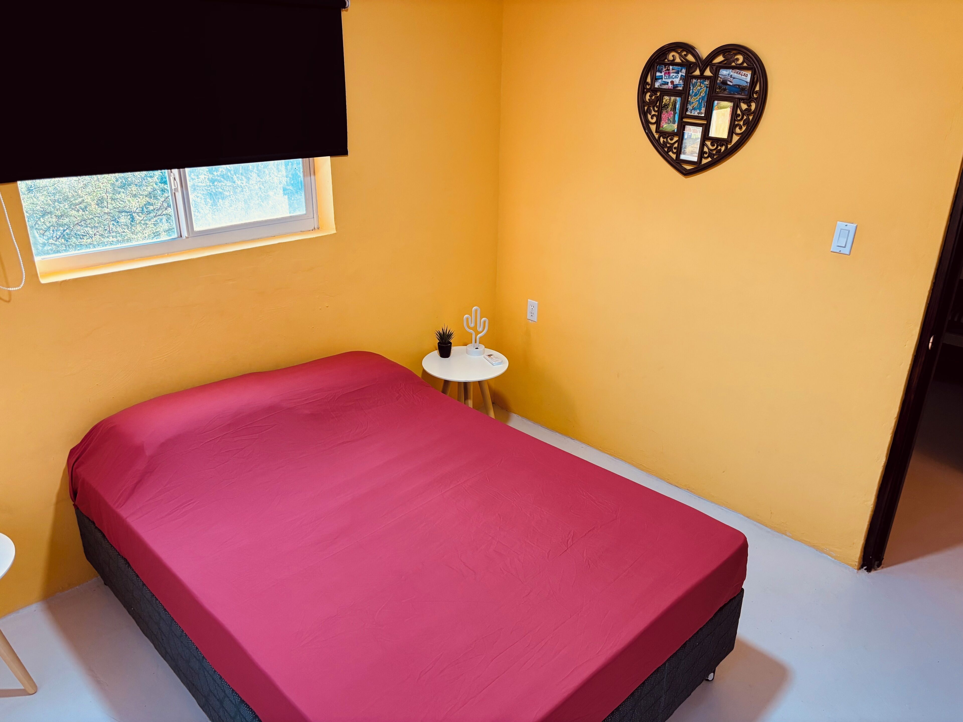 Una habitación con una cama, una mesita con un cactus, un reloj de pared y una ventana.
