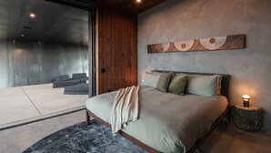 A modern bedroom with a large bed, a wooden headboard, a wall-mounted abstract art piece, and a bedside lamp. - Obsidian - Industrial-Modern Villa (Yucca Valley)
