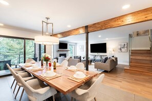 A modern dining area with a wooden table set for four, surrounded by comfortable chairs, a chandelier, and a large window offering a view of greenery.