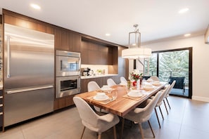 Een moderne keuken met een roestvrijstalen koelkast, oven en een inbouwmagnetron. Er is een eettafel met stoelen, een kroonluchter en een uitzicht op het buitenleven door een groot raam.