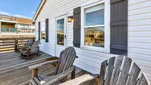 A wooden deck with Adirondack chairs, a white building with large windows, and a view of a dock and boats.