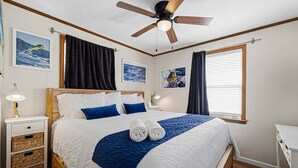 A neatly made bed with a blue blanket and two rolled towels, a ceiling fan, and framed pictures on the wall.