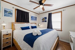 A neatly made bed with a blue blanket and two rolled towels, a ceiling fan, and framed pictures on the wall. - Billfish Bungalow | 1950 ft to the beach | Dog Friendly and Hot Tub (Nags Head)
