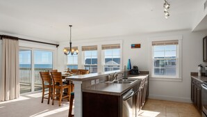 Una cocina moderna con una isla grande, un comedor con sillas y vistas al océano a través de amplios ventanales.