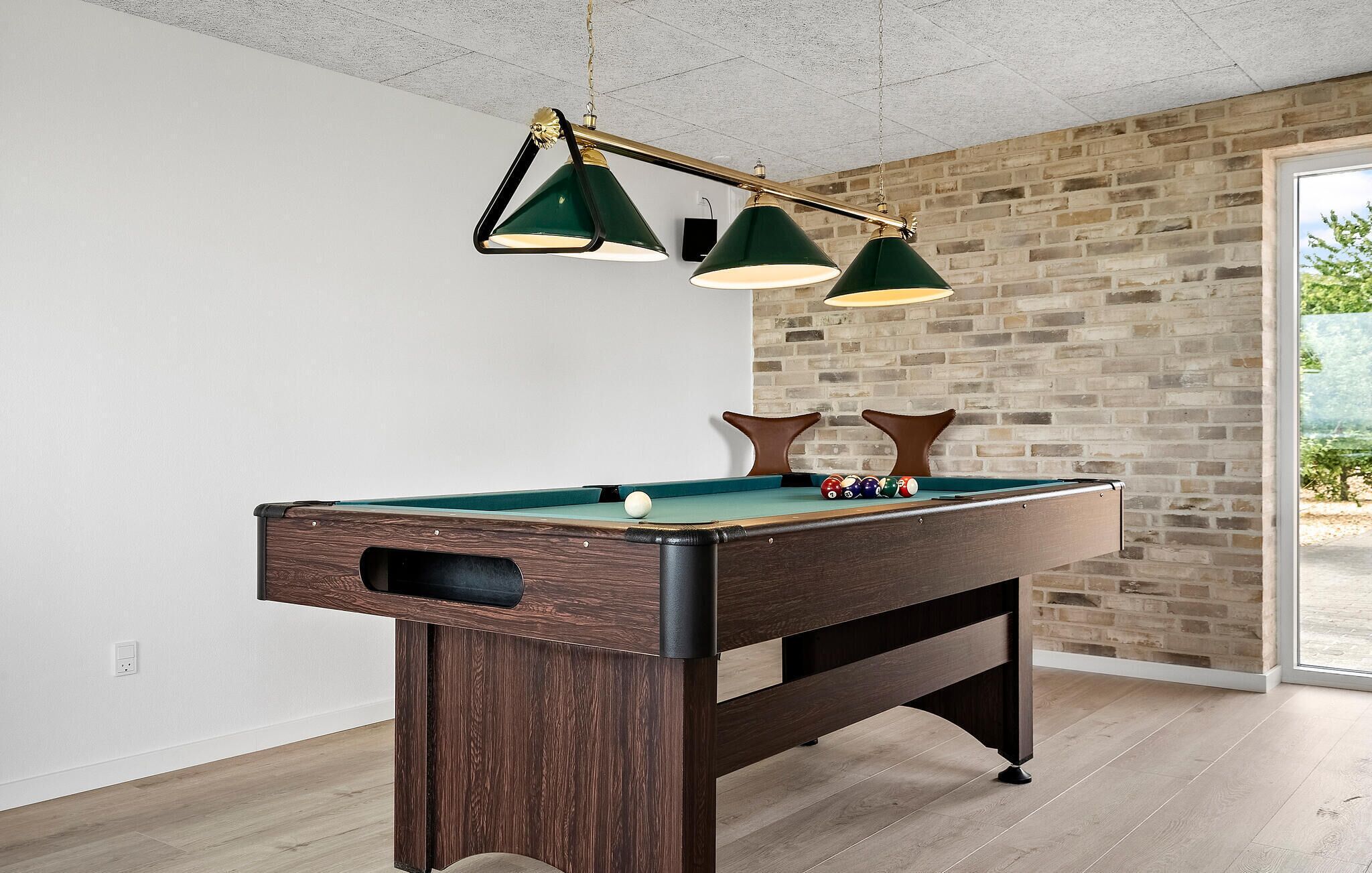 A pool table with a wooden finish, three green pendant lights, and a brick wall in the background.