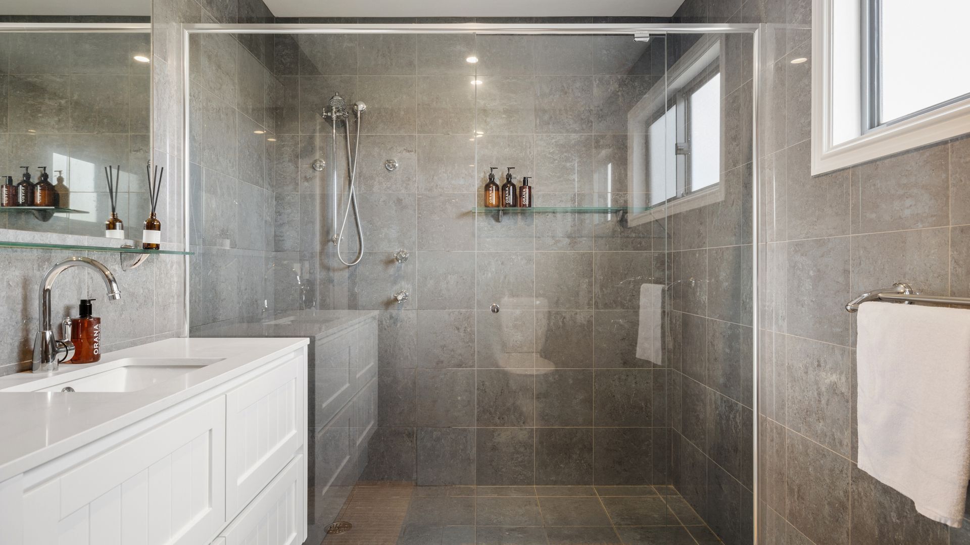 Un baño moderno con ducha de vidrio, tocador blanco y repisa de vidrio con artículos de aseo.