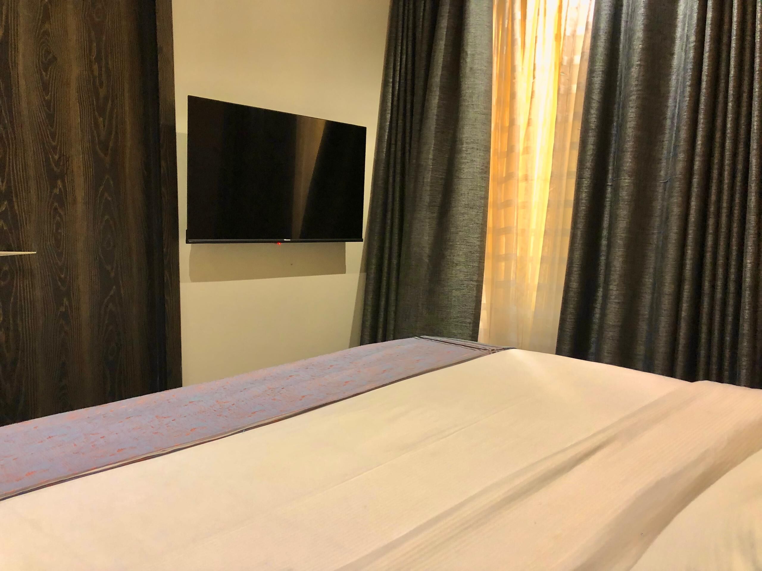 A hotel room with a bed, a flat-screen TV mounted on the wall, and dark curtains.