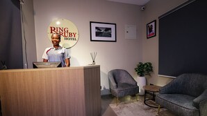 Un hall d’hôtel avec un comptoir d’accueil en bois, une grande enseigne indiquant « Ring Ruby Hotel », une photo encadrée accrochée au mur et deux fauteuils gris.