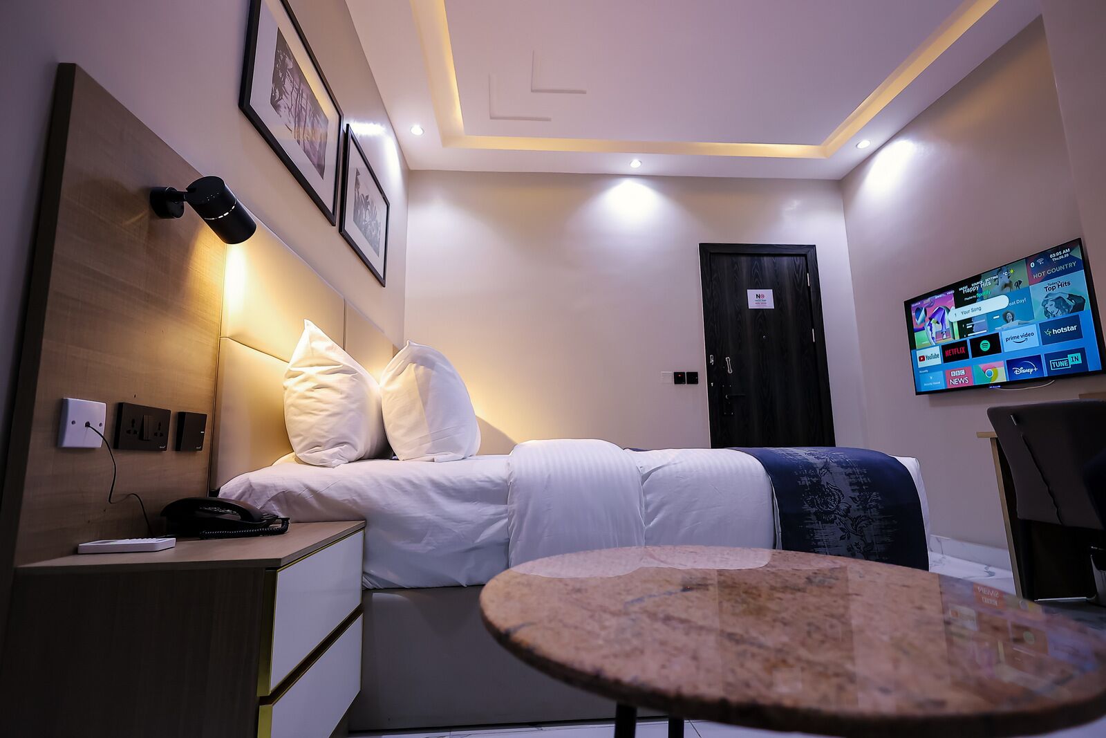 A hotel room with a bed, bedside table, lamp, wall-mounted TV, and a small round table.