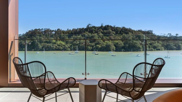 Un balcon avec deux chaises donnant sur une baie où se trouvent des bateaux et une colline boisée.