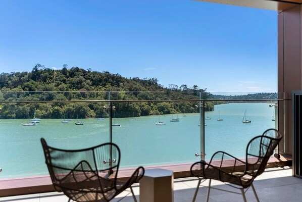 A balcony with two chairs overlooking a bay with boats, a forested hillside, and a clear sky.