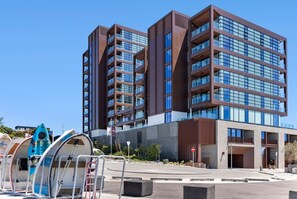A modern multi-story building with a mix of glass and metal facade, a parking area, and a blue boat sculpture.