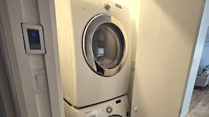 A compact laundry room with a Samsung washer and dryer, a control panel, and a small storage shelf.