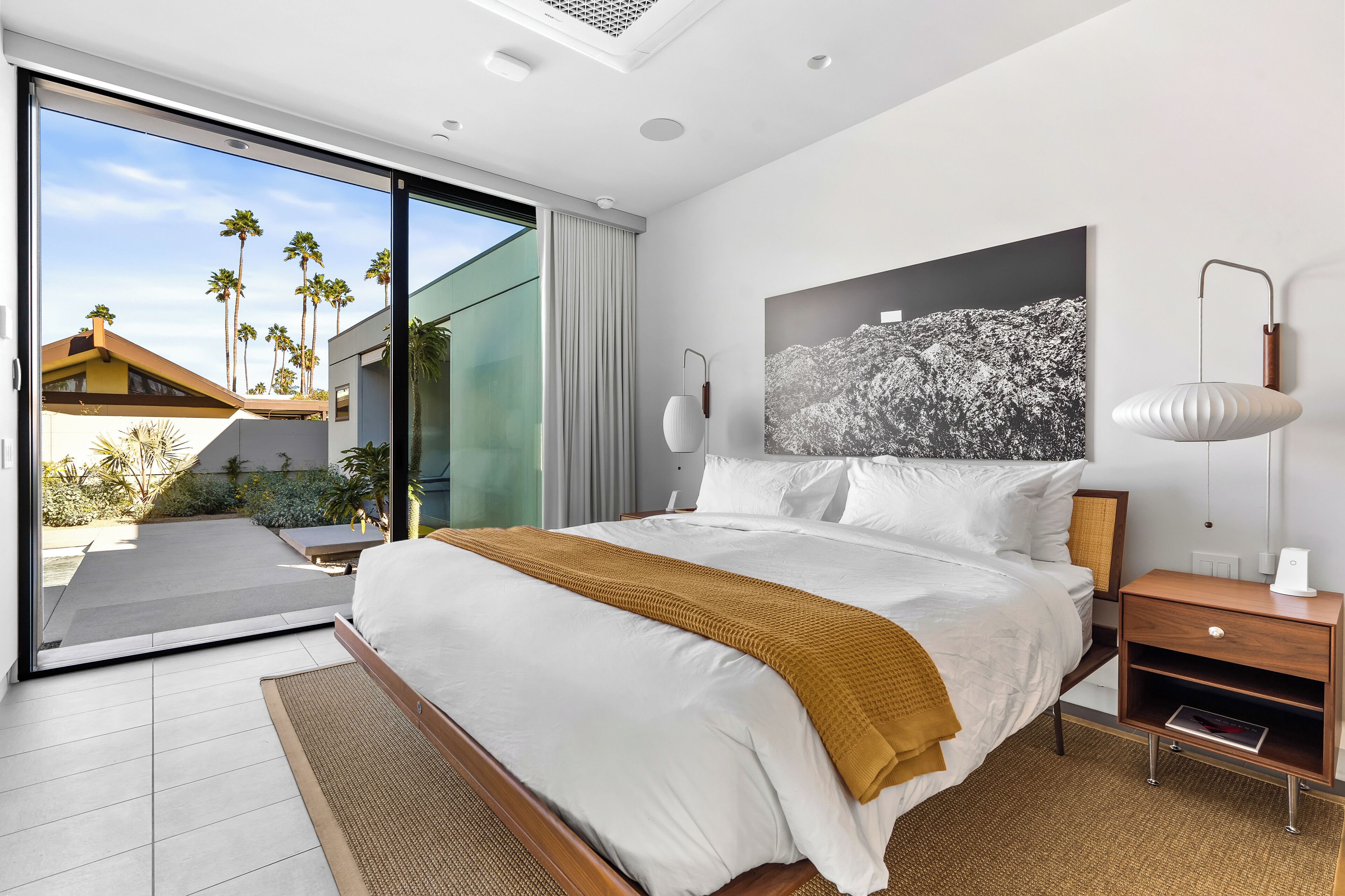 A modern hotel room with a large bed, a nightstand, a lamp, and a view of palm trees through a sliding glass door.