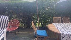 An outdoor patio area with a wooden table, a striped lounge chair, a red woven chair, and a blue cushioned chair under a large umbrella.
