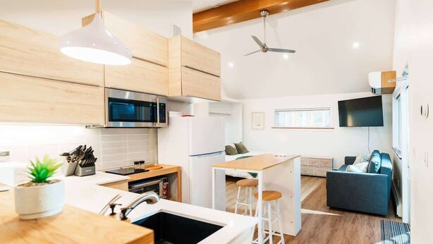 Cozinha moderna com armários de madeira, micro-ondas e pia. Há uma área de jantar com mesa e cadeiras, e uma TV fixada na parede.