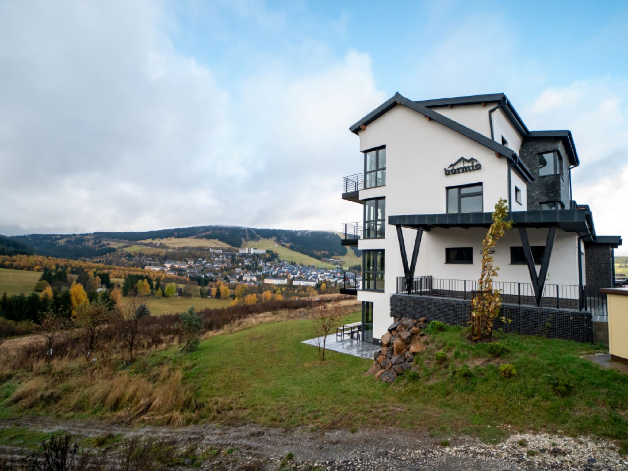 A modern two-story hotel with a balcony, set against a backdrop of a rural landscape and distant town.
