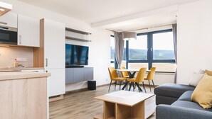 Ein modernes Wohnzimmer mit einer Couch, einem Couchtisch und einem Essbereich mit Stühlen. Es gibt eine Küchenzeile mit Schränken und einem Einbauofen. Ein großes Fenster bietet einen Blick ins Freie.