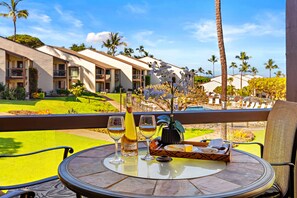 Ein Tisch mit einer Champagnerflasche, zwei Gläsern, einer Brottafel und einem Blumenstrauß auf einem Balkon mit Blick auf einen Pool und Hotelgebäude.
