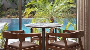 A round table with four wooden chairs and a glass of water, set against a backdrop of a swimming pool and tropical plants.