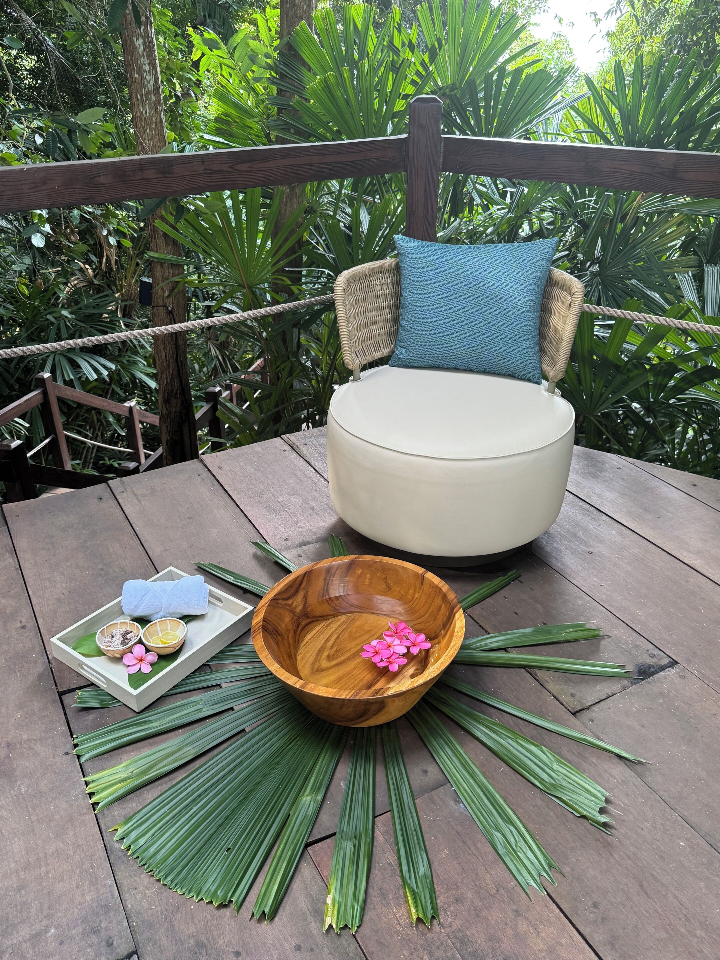 A wooden deck with a round white chair, a woven chair with a blue cushion, and a wooden bowl with pink flowers.