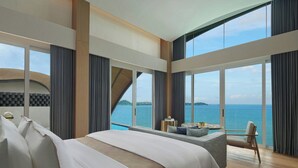 Una habitación de hotel moderna con una cama grande, una zona de estar con vistas al mar y ventanas amplias.