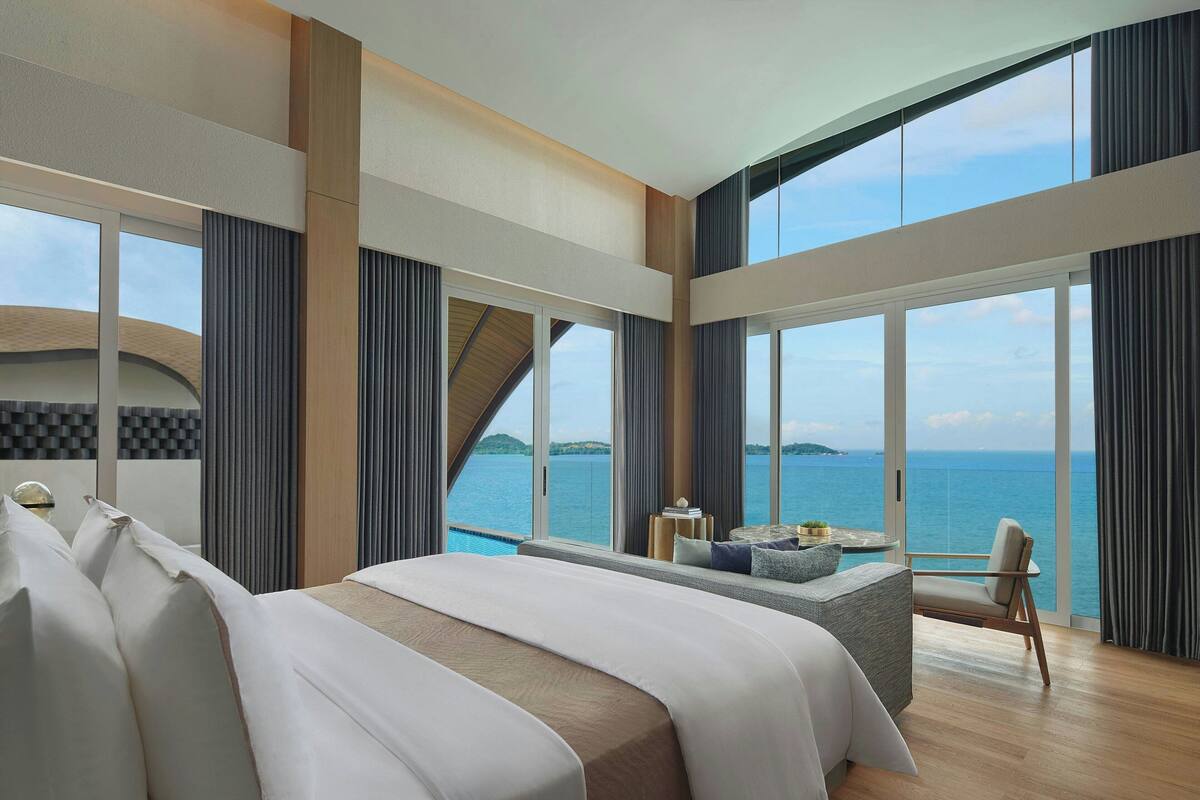 Una habitación de hotel moderna con una cama grande, una zona de estar con vistas al mar y ventanas amplias.