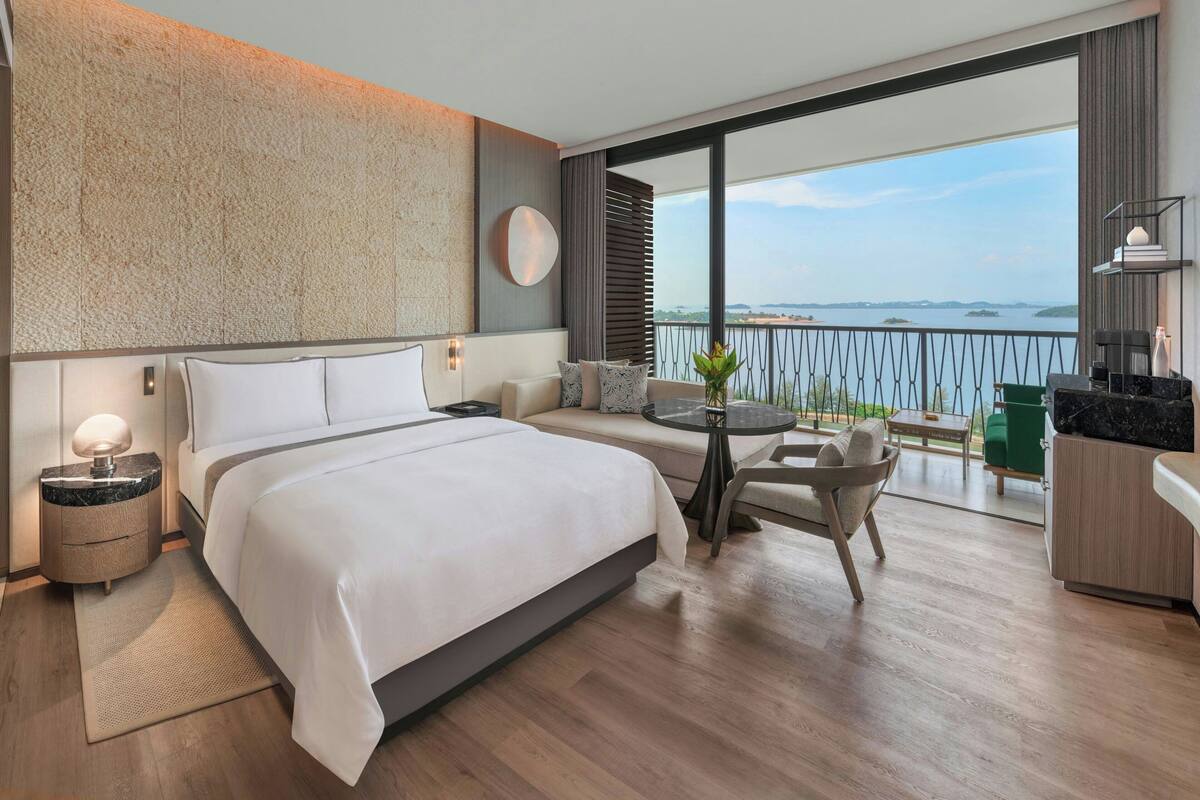 Una habitación de hotel moderna con una cama grande, una pequeña mesa redonda, un sofá y un balcón con vista al mar.