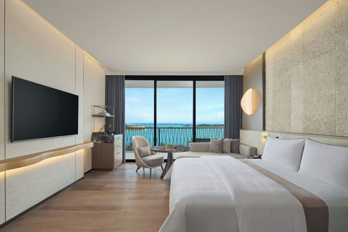 Una habitación de hotel moderna con una cama grande, un televisor de pantalla plana, un balcón con vistas al mar y una zona de estar con una mesa y sillas.