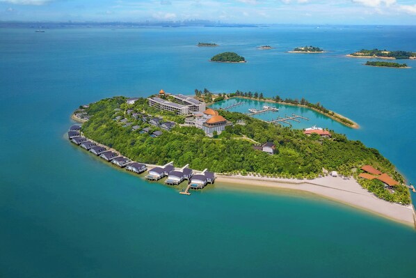 Vista aérea de um resort em uma ilha tropical, com um edifício central rodeado por vegetação exuberante, uma marina e uma praia.