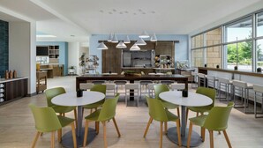 A modern kitchen with a central island, dining area with round tables and green chairs, and large windows offering a view of the outdoors. - Element by Marriott Charleston Airport (North Charleston)