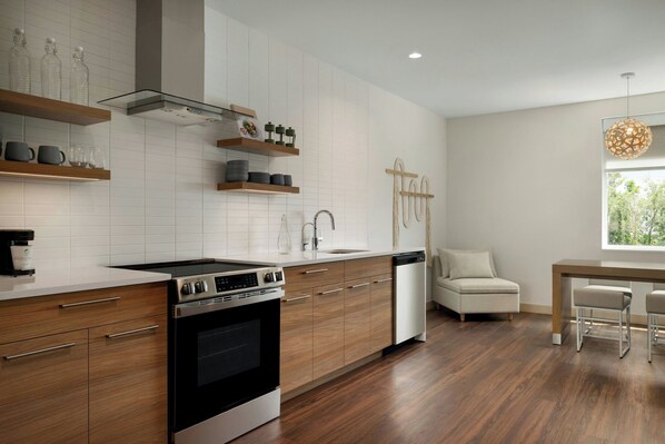A modern kitchen with wooden cabinets, a built-in oven, and a dishwasher. There is a dining area with a table and chairs. - Element by Marriott Charleston Airport (North Charleston)