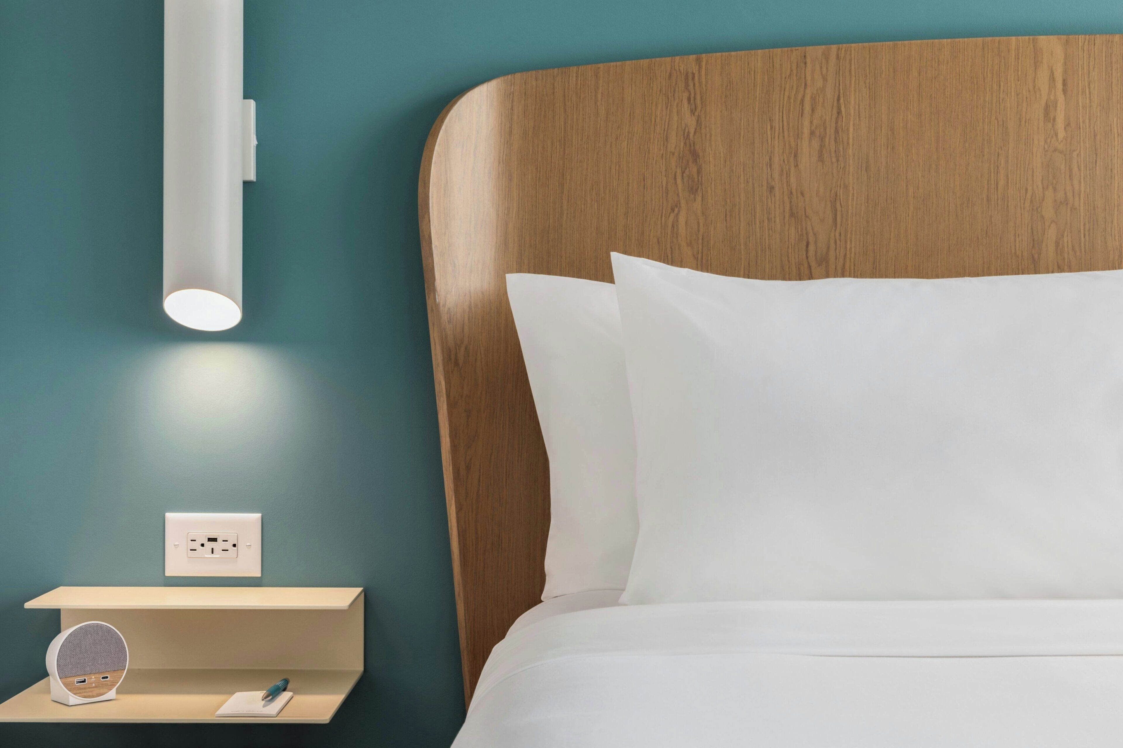 Una cama bien hecha con sábanas blancas, un cabecero de madera, una mesita de noche con lámpara y un enchufe.