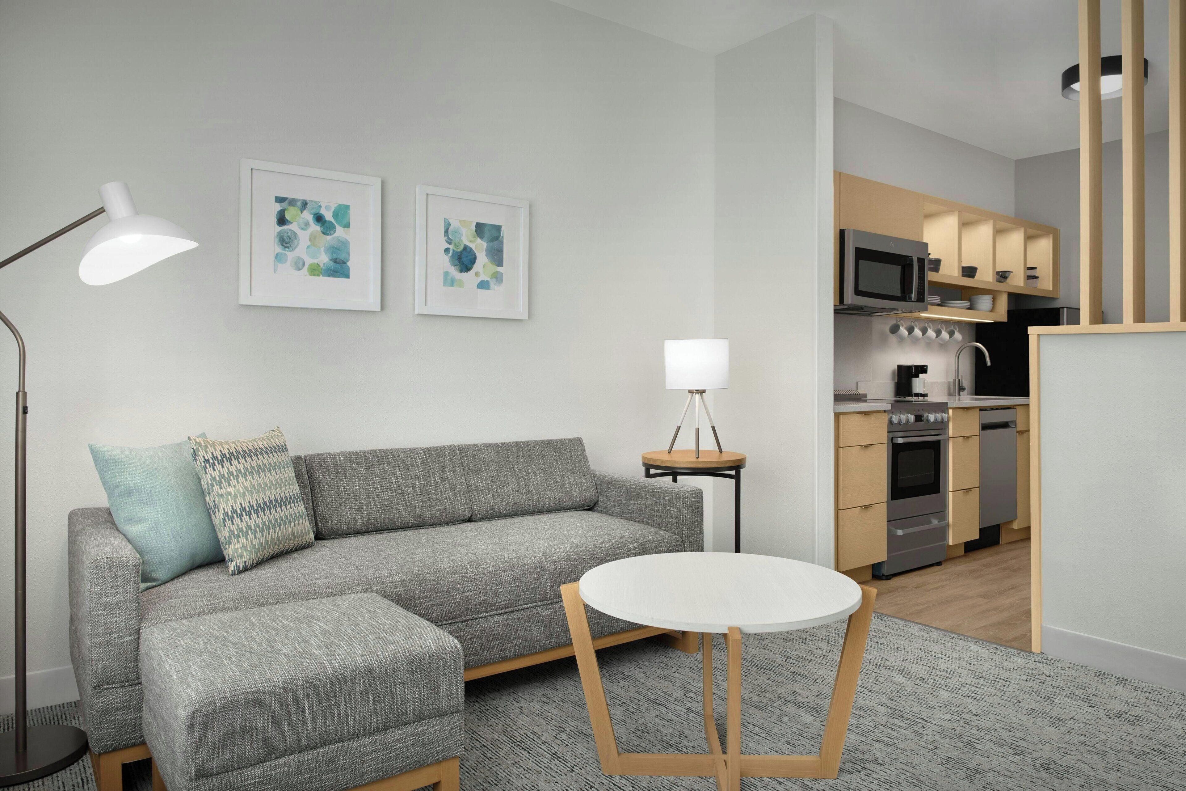 A modern living room with a grey sofa, a round table, and a kitchen area with built-in appliances.