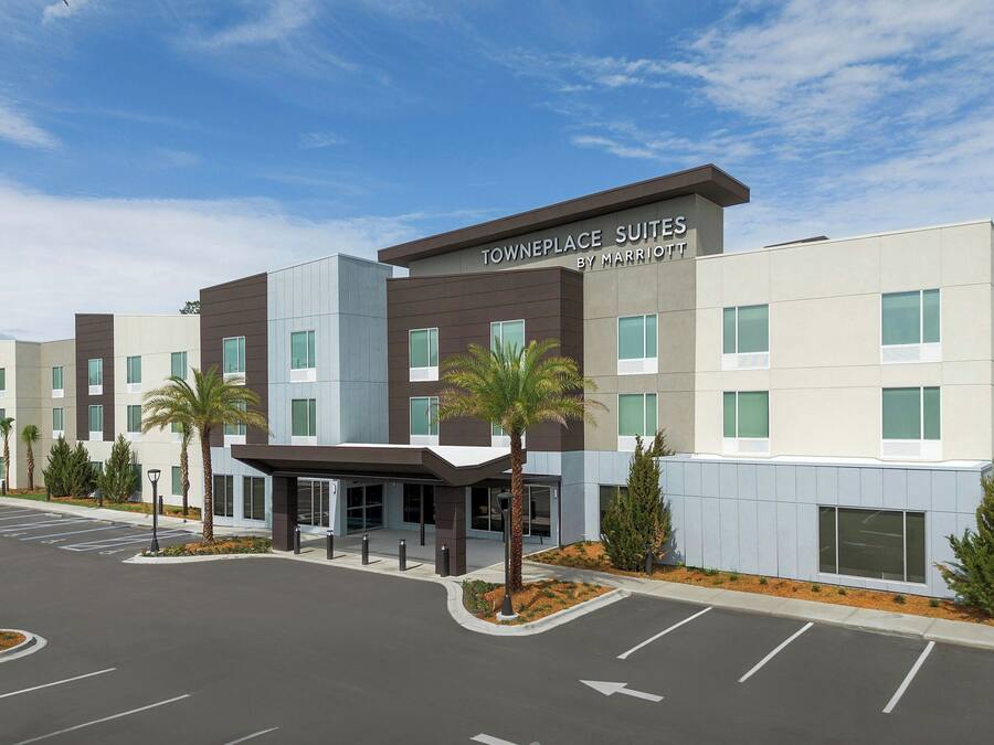 A modern hotel with a sign reading "Towneplace Suites by Marriott" featuring a covered entrance, parking lot, and landscaping with palm trees.
