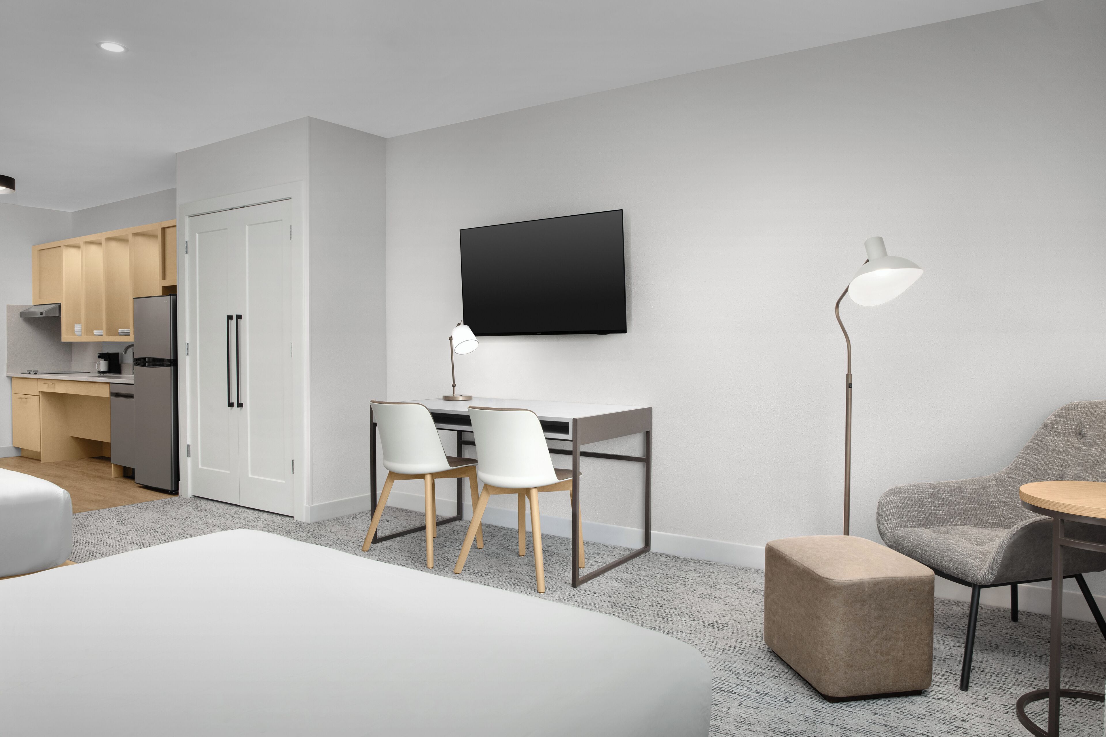 Una habitación de hotel moderna con una cama, un escritorio con dos sillas, un televisor de pantalla plana, una lámpara y una kitchenette con armarios y electrodomésticos.